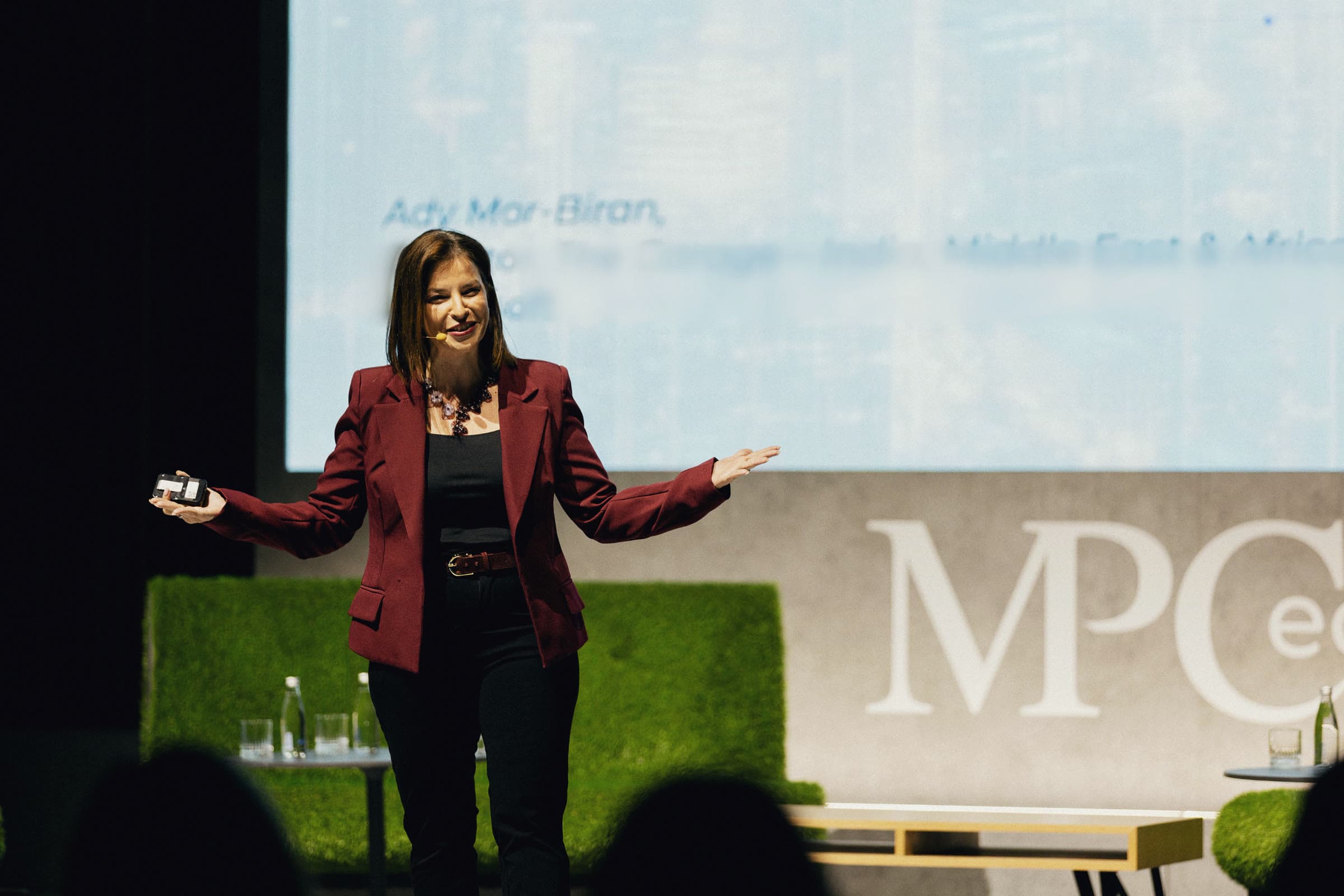 Ady Mor-Biran on a conference stage in a burgundy blazer, arms open toward the audience, a large screen behind her and audience silhouettes in the foreground.